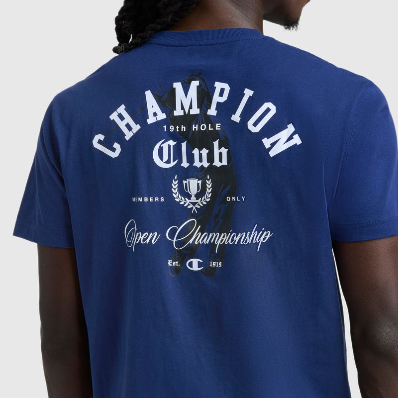 A person wears the Champion Club Graphic T-Shirt by Champion, a soft cotton jersey tee with a C logo and white back text: “CHAMPION Club 19th HOLE, Open Championship, Est. 1919,” plus graphic details and laurel leaves.
