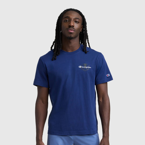 A person with long, dark braided hair wears a blue Champion Club Graphic T-Shirt, C Logo by Champion, made from soft cotton jersey, standing against a plain light gray background.