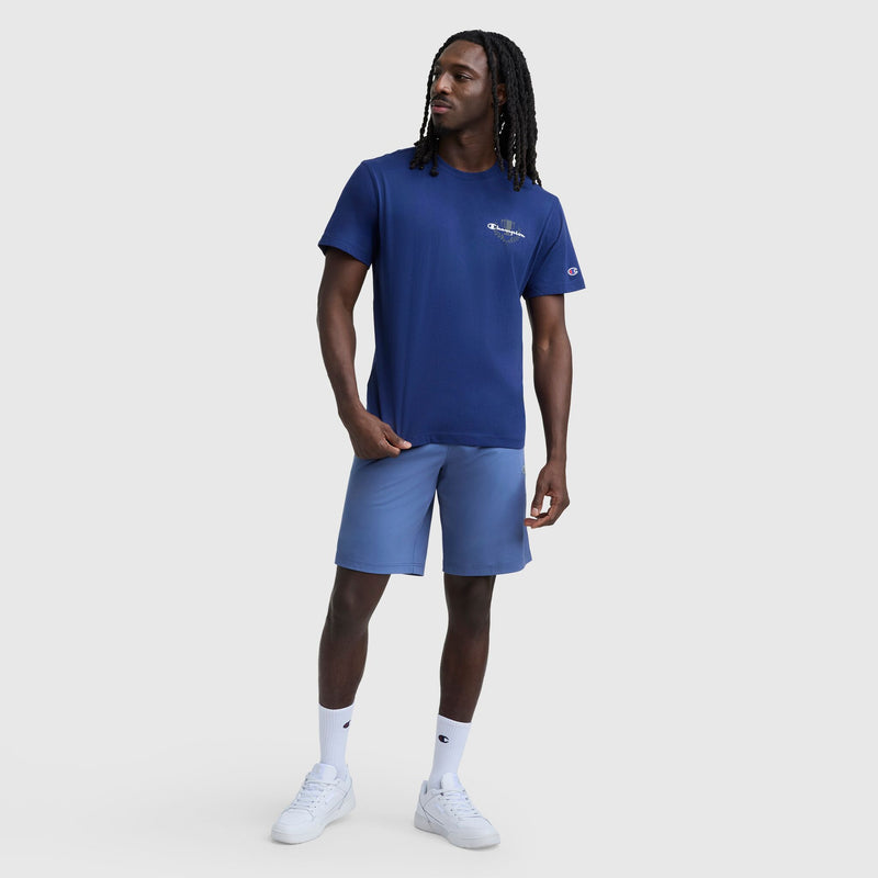 A man with long braids wears a navy blue Champion Club Graphic T-Shirt, C Logo by Champion, paired with light blue shorts, white crew socks, and white sneakers, standing against a plain light gray background.