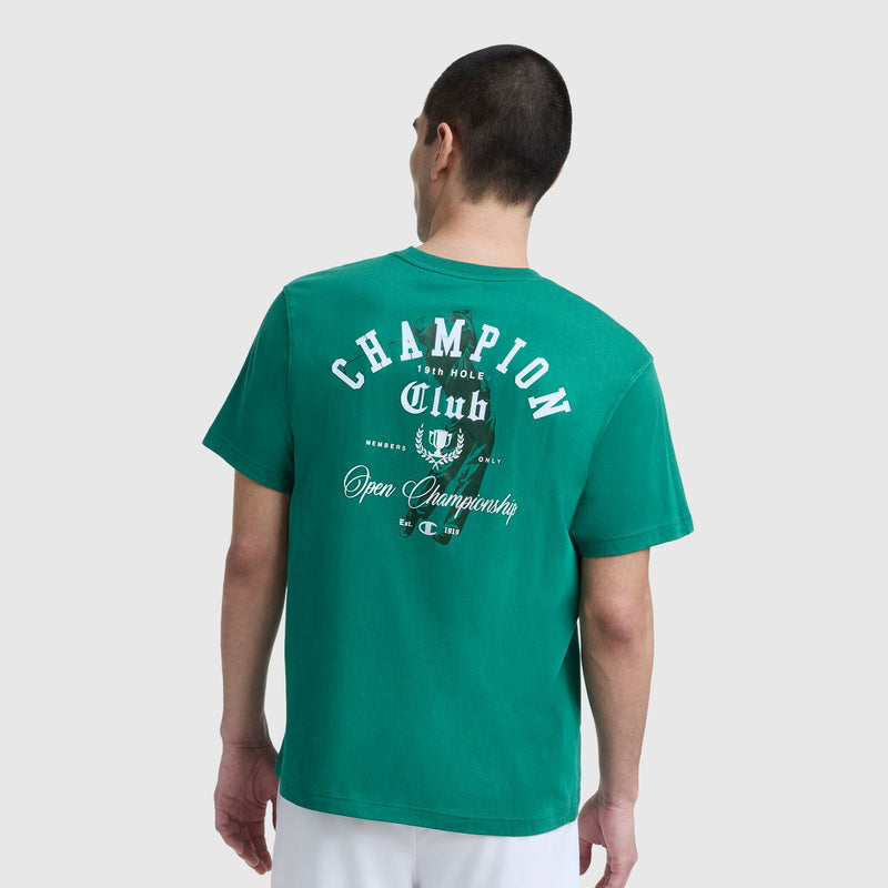 A person wears a green Champion Club Graphic T-Shirt, C Logo by Champion, with white "CHAMPION Club Open Championship" graphics on the back, shown from behind against a plain light background.