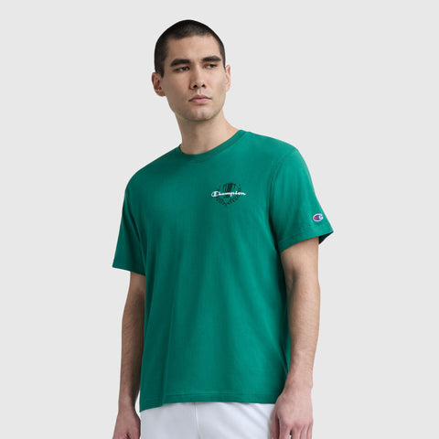 A man with short dark hair wears a green Champion Club Graphic T-Shirt, C Logo by Champion, made of soft cotton jersey with a small white logo on the chest, paired with white pants against a plain light gray background.