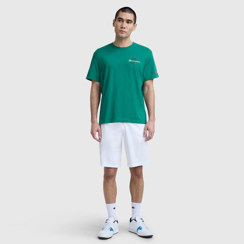 A man stands against a light gray background, wearing a green Champion Club Graphic T-Shirt with C Logo, white knee-length shorts, white crew socks, and white sneakers featuring blue accents.