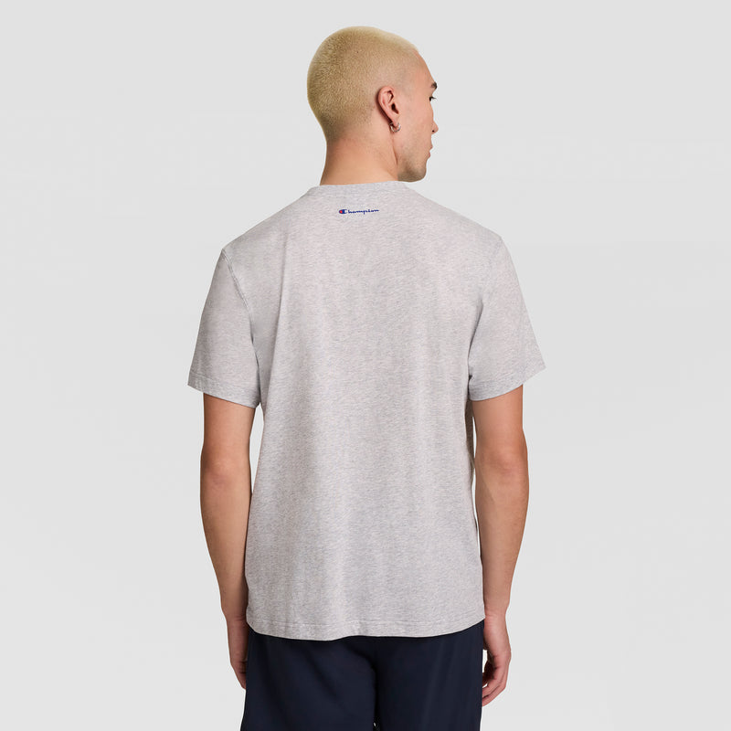 A person with short blond hair is shown from behind, wearing a light gray Champion Short Sleeve USA T-Shirt and dark shorts, standing against a plain light background.