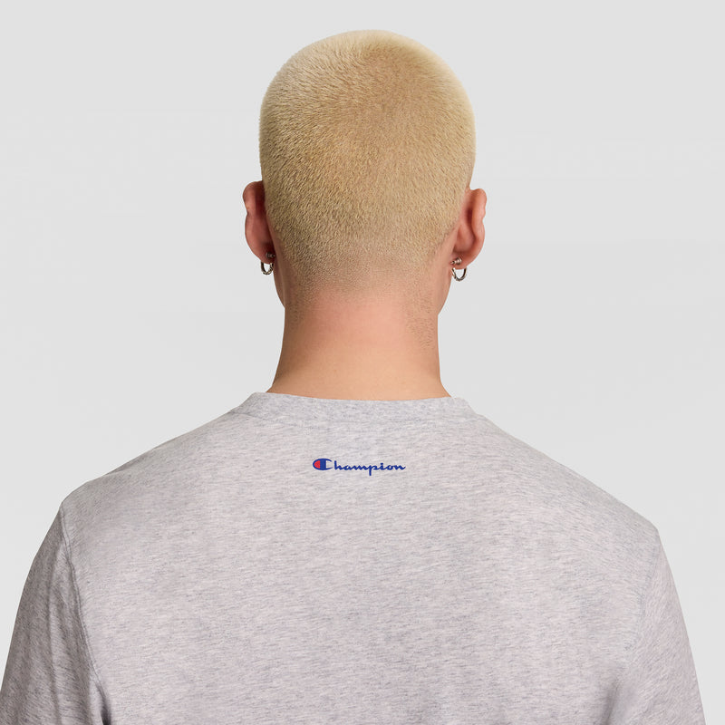 A person with short platinum blonde hair and silver hoop earrings is seen from behind against a plain light background, wearing the Champion Short Sleeve USA T-Shirt.