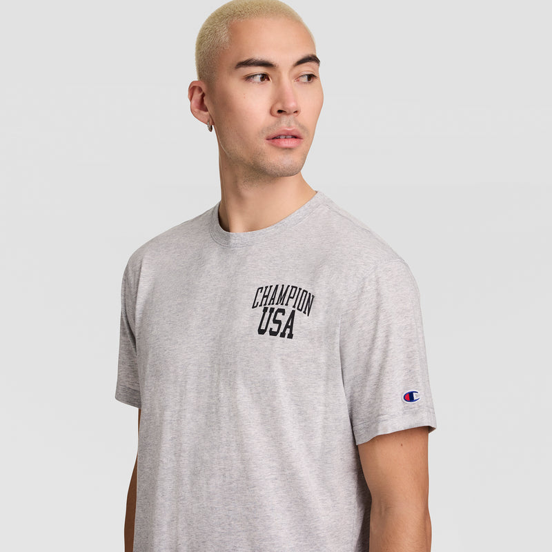A person with short blond hair wears a gray Champion Short Sleeve USA T-Shirt and looks to their left. The athletic shirt features a small Champion logo on the sleeve and “CHAMPION USA” printed across the chest.