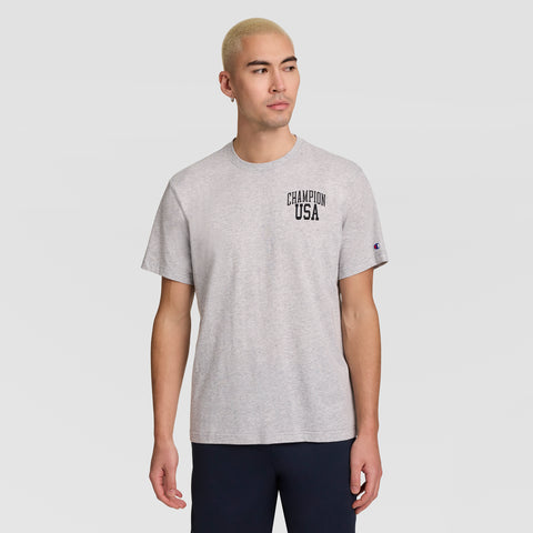A man with short blond hair wears a Champion Short Sleeve USA T-Shirt in gray and dark pants, standing against a plain light gray background while looking to his left.
