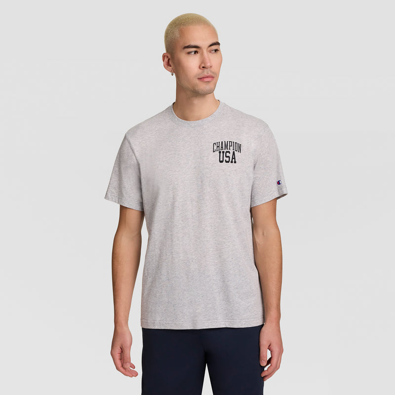 A man with short blond hair wears a Champion Short Sleeve USA T-Shirt in gray and dark pants, standing against a plain light gray background while looking to his left.