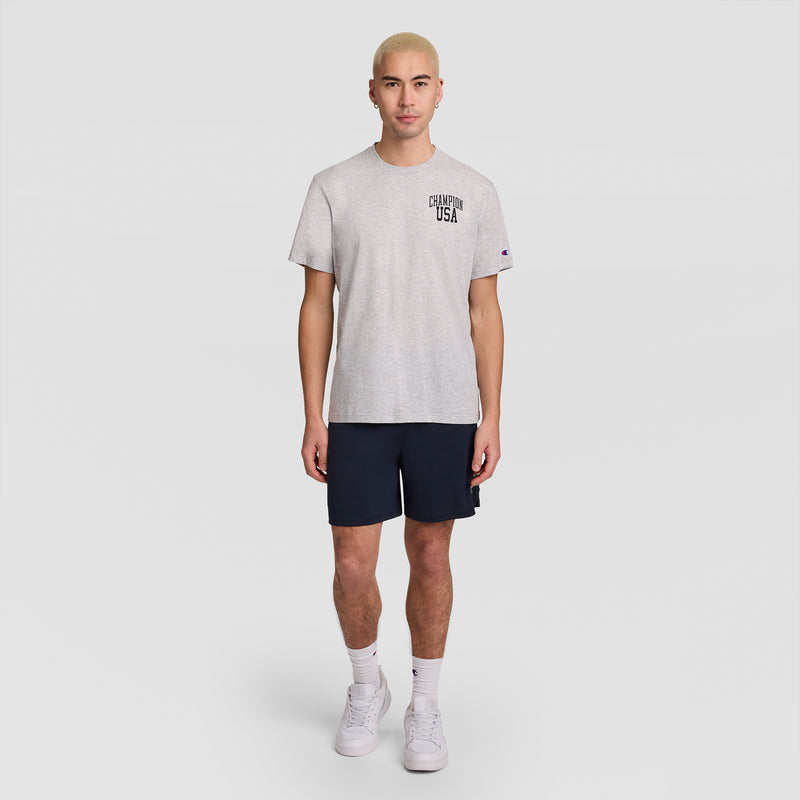 A person with short blonde hair wears a Champion Short Sleeve USA T-Shirt in gray, paired with navy shorts, white socks, and white sneakers, standing against a plain light gray background.