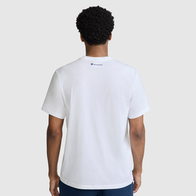 A person is shown from the back, wearing the Champion Short Sleeve USA T-Shirt in plain white with a small Champion logo on the upper back, paired with navy blue pants. The background is light gray.