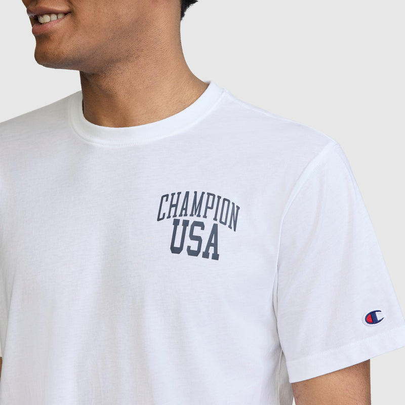 A person wears the Champion Short Sleeve USA T-Shirt—a white graphic tee featuring "CHAMPION USA" in bold letters across the chest and a small Champion logo patch on the left sleeve. The casual, athletic shirt is styled simply as the person smiles, turned slightly to the left.