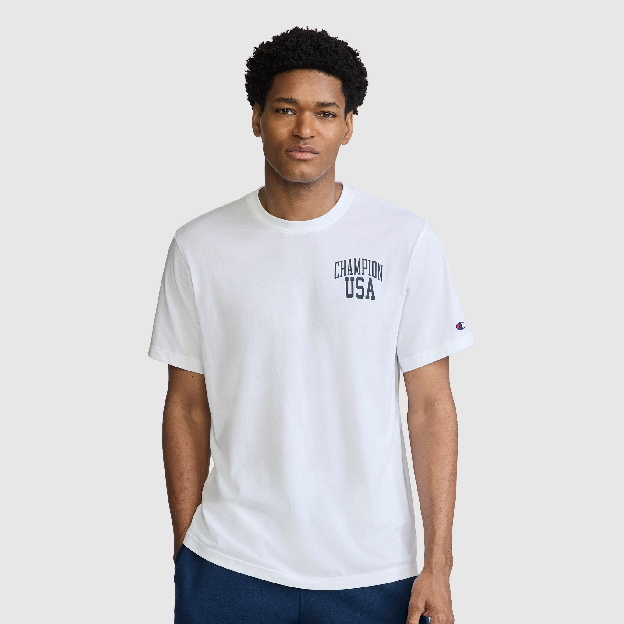 A man wearing a Champion Short Sleeve USA T-Shirt, featuring "Champion USA" printed on the chest, stands against a plain light gray background, showcasing this classic athletic piece.