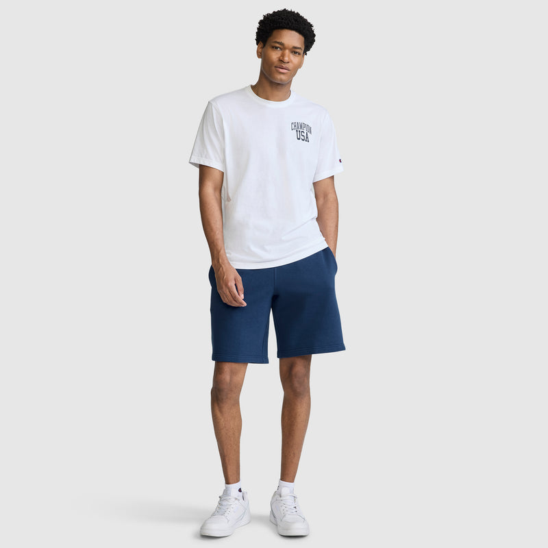 A person stands facing forward, wearing the Champion Short Sleeve USA T-Shirt—a classic white athletic tee featuring "USA" across the chest—paired with navy blue shorts and white sneakers. The background is plain and light gray.
