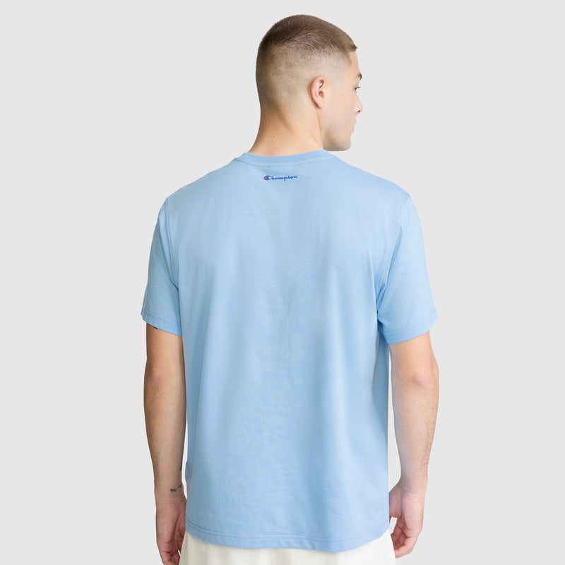 A man with short hair, dressed in a light blue Champion Short Sleeve USA T-Shirt and white shorts, stands with his back to the camera against a plain gray background.