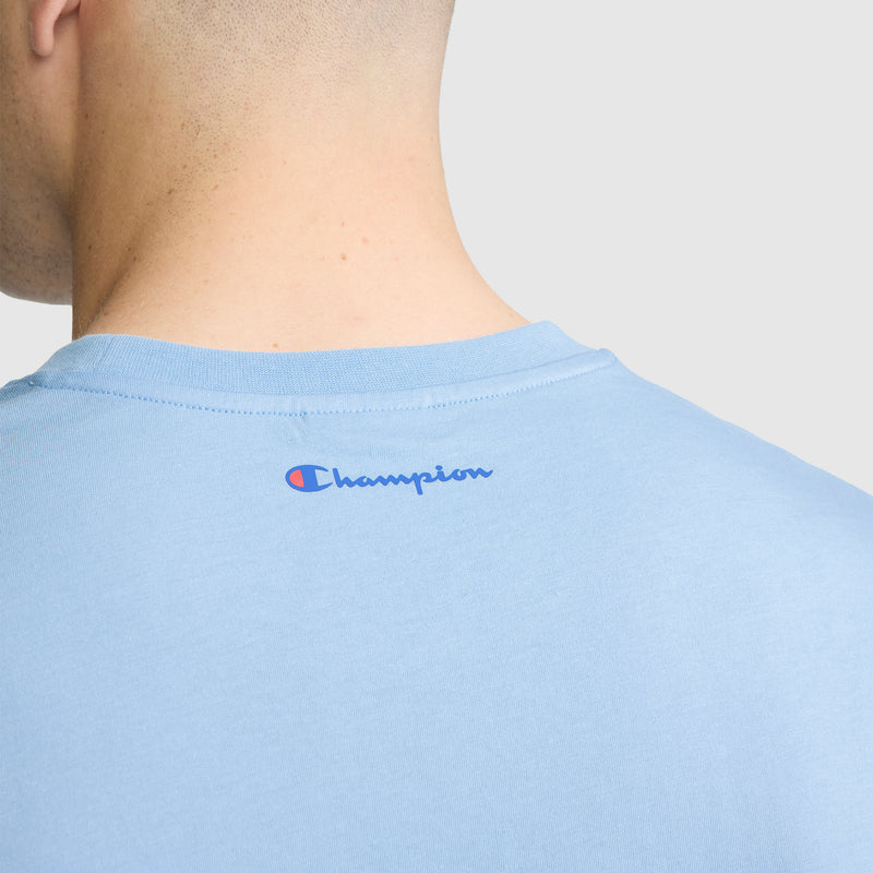 Close-up of the back of a person's neck and shoulders wearing a light blue Short Sleeve USA T-Shirt by Champion, featuring the brand's logo in blue near the collar. This classic men’s tee offers Champion’s signature comfort and timeless sporty style.