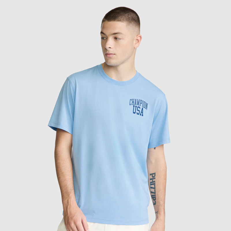 A young man stands against a plain light gray background, wearing a Champion Short Sleeve USA T-Shirt in light blue. He looks to his left with his right hand partially in his pocket.