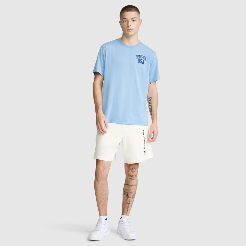 A young man stands facing forward wearing a Champion Short Sleeve USA T-Shirt in light blue, paired with off-white shorts and white sneakers. He has visible tattoos on his left arm and right leg. The background is plain and light gray.