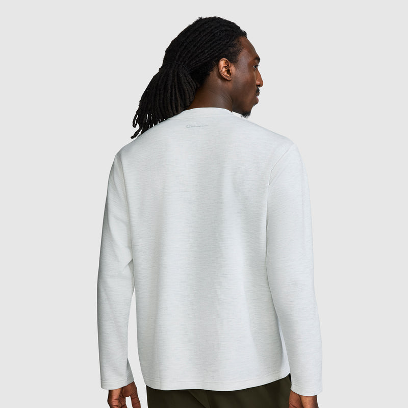 A person with long dark hair styled in dreadlocks is wearing a light gray Champion Sport Long Sleeve Crewneck, C Logo shirt and dark pants, standing against a plain light background, facing away from the camera.