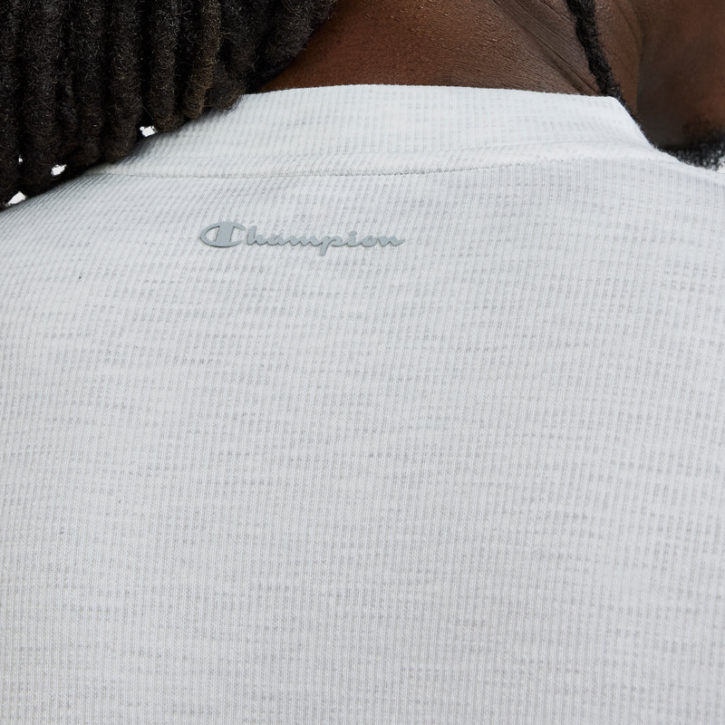 Close-up of a person’s shoulder and neck wearing a white textured Champion Sport Long Sleeve Crewneck, C Logo performance top with the Champion logo printed on the back. The person’s hair is styled in short dreadlocks.