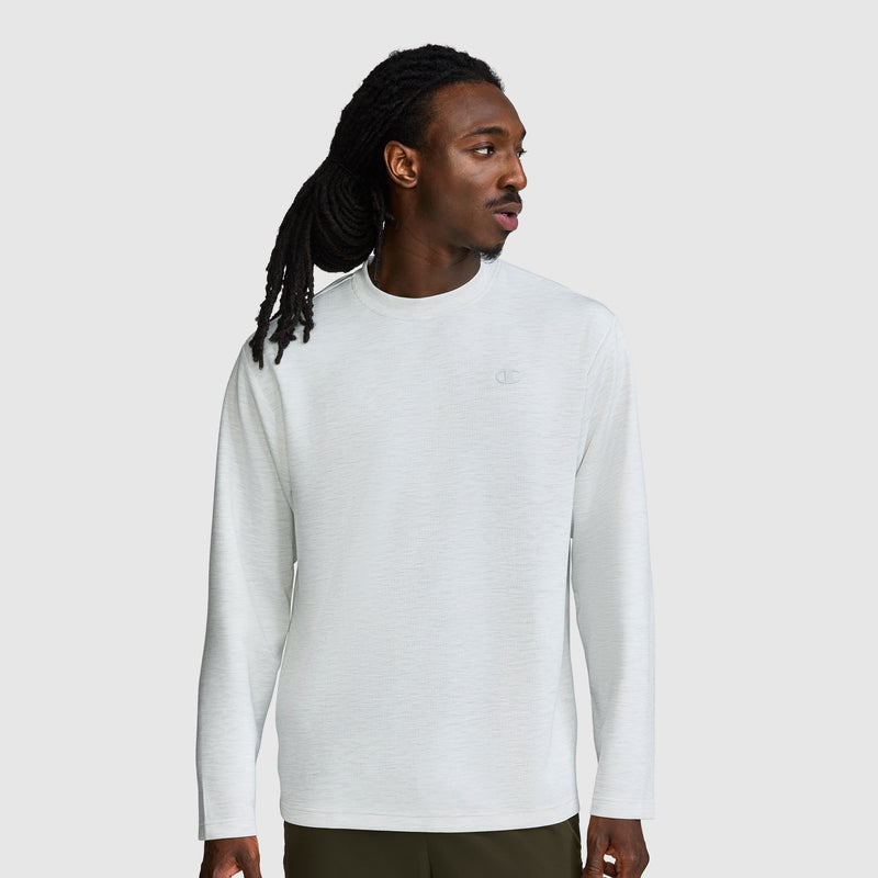 A man with long dreadlocks wears a Champion Sport Long Sleeve Crewneck, C Logo in light gray and dark pants, standing against a plain light gray background while looking to his left.