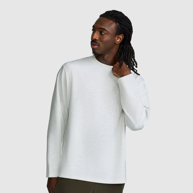 A man in a light gray Champion Sport Long Sleeve Crewneck, featuring the C Logo, stands against a plain light background, looking to the side with one hand touching the back of his neck.