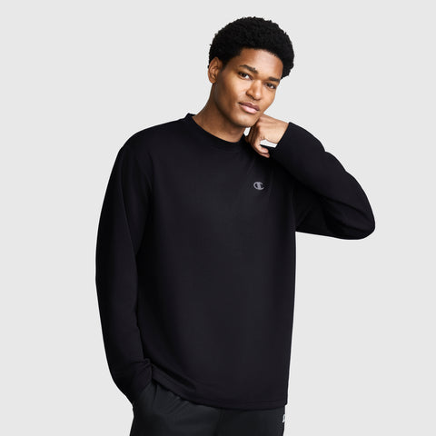 A person wearing a loose-fitting black Champion Sport Long Sleeve Crewneck with a small C logo on the chest stands against a plain light gray background, looking at the camera with one hand touching their neck.