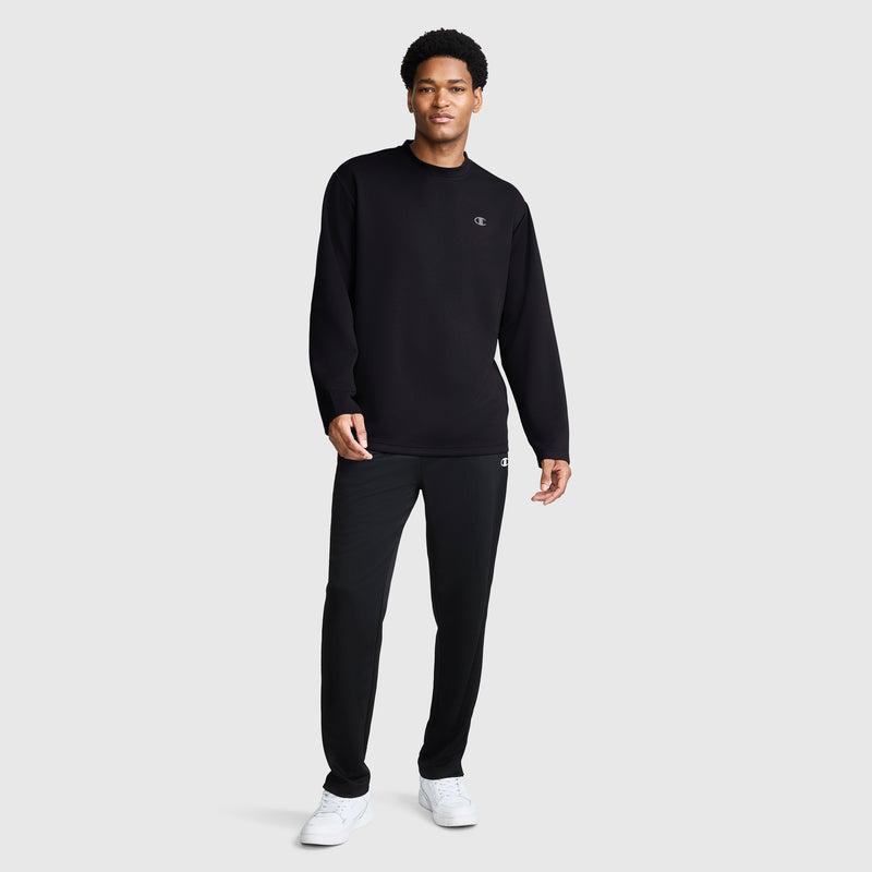 A man stands facing forward, wearing a black Champion Sport Long Sleeve Crewneck, C Logo, paired with matching black pants and white sneakers. The plain light gray background highlights his modern athletic wear ensemble.
