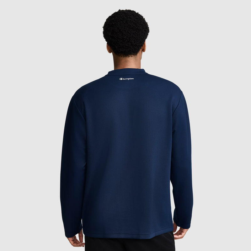 A person stands facing away, wearing a navy blue Champion Sport Long Sleeve Crewneck C Logo shirt and black pants. The background is plain light gray.