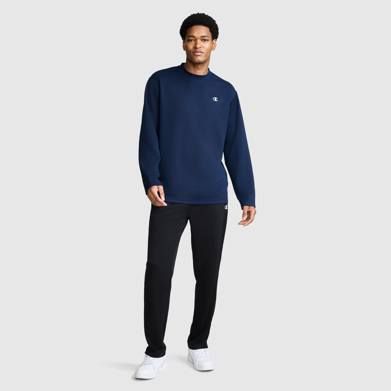 A man stands against a plain background wearing a navy blue Champion Sport Long Sleeve Crewneck, C Logo top, black pants, and white sneakers.