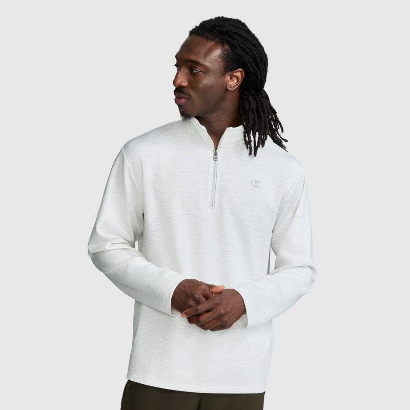 A man with long dreadlocks wears a light gray Champion Sport Long Sleeve 1/4 Zip Mockneck with the C Logo and dark pants, standing against a plain light background while looking to his left.