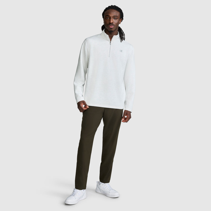 A man stands against a plain background wearing a light gray Champion Sport Long Sleeve 1/4 Zip Mockneck with the C Logo, paired with dark pants and white sneakers. His long hair is styled in twists, and he looks at the camera with a neutral expression.