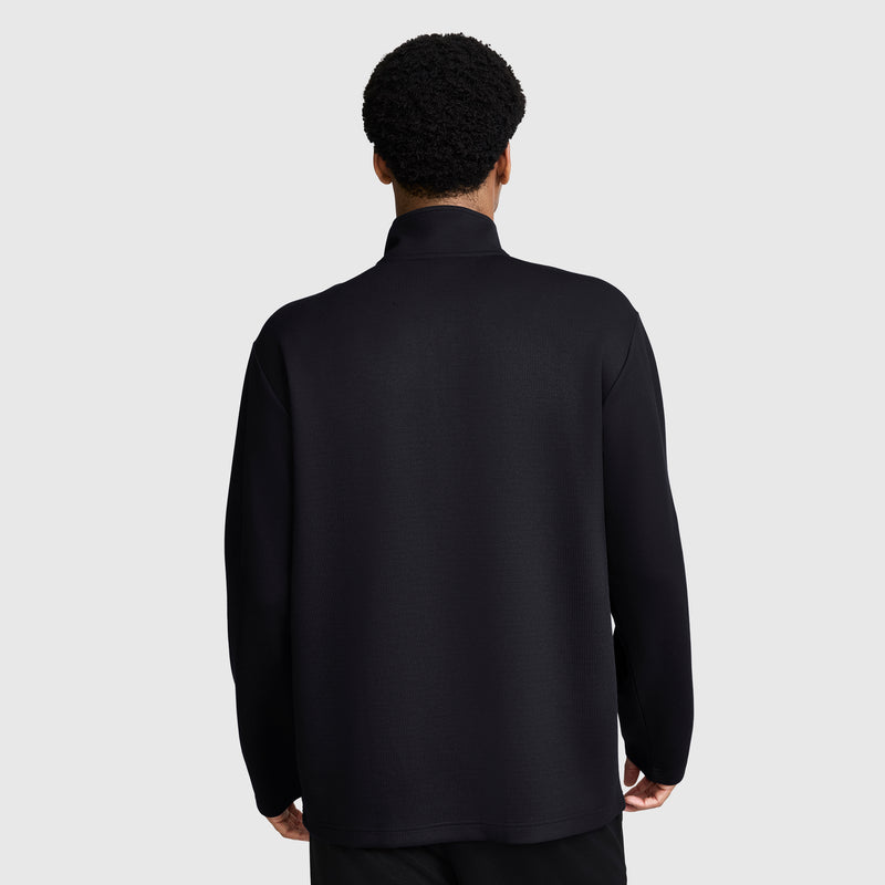 A person with short curly hair is seen from behind, wearing a Champion Sport Long Sleeve 1/4 Zip Mockneck with the C Logo and black pants, standing against a plain light gray background.
