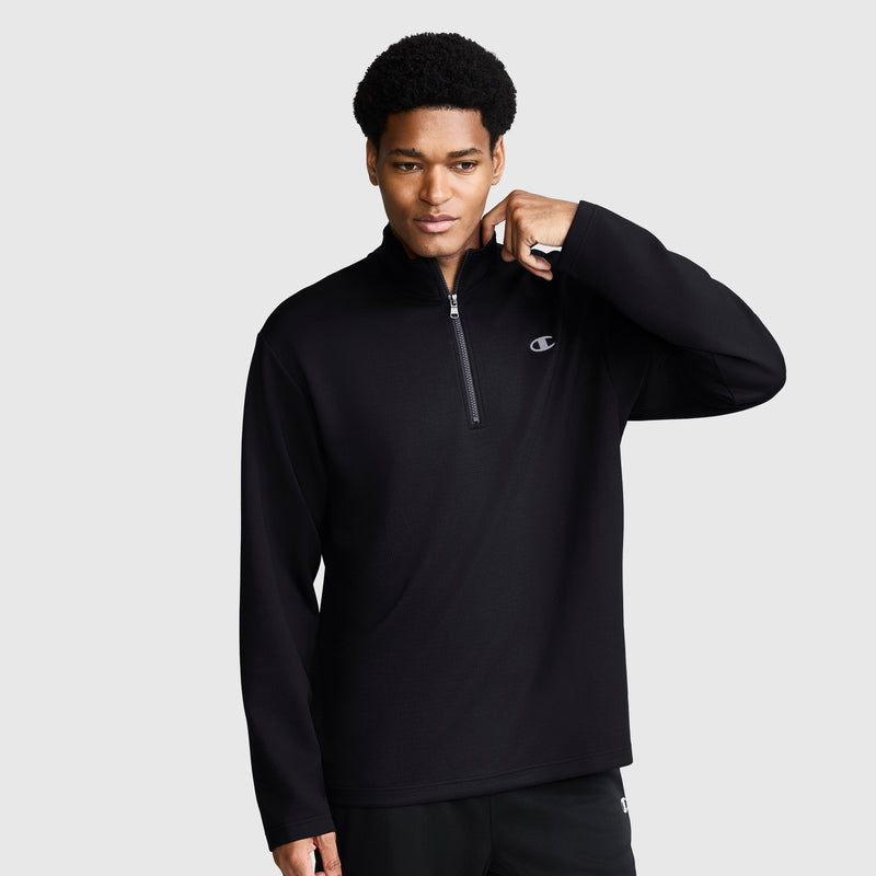 A man wears the Champion Sport Long Sleeve 1/4 Zip Mockneck featuring the signature C Logo in black, standing against a plain light gray background while holding the collar with one hand and gazing slightly to the side.