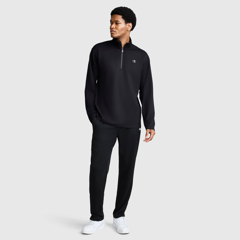 A man stands wearing the Champion Sport Long Sleeve 1/4 Zip Mockneck with C Logo and matching black pants, paired with white sneakers. He is looking to his left, left hand by his side and right hand relaxed, set against a plain light gray background that highlights the sleek style of this men's quarter zip pullover.