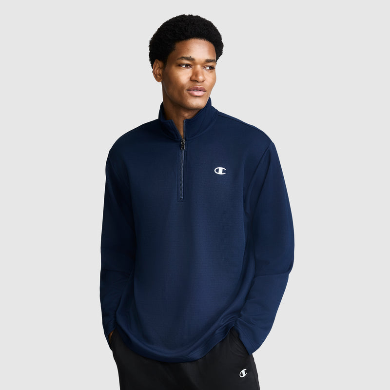 A man stands against a plain light gray background with his hands in his pockets, wearing black pants and a Champion Sport Long Sleeve 1/4 Zip Mockneck pullover featuring the C Logo in navy blue, looking off to the side.