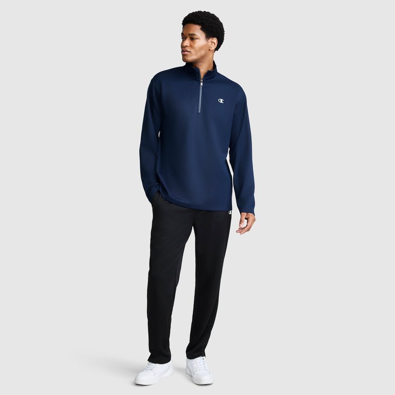 A man stands against a plain light gray background wearing the Champion Sport Long Sleeve 1/4 Zip Mockneck with C Logo, paired with black pants and white sneakers.