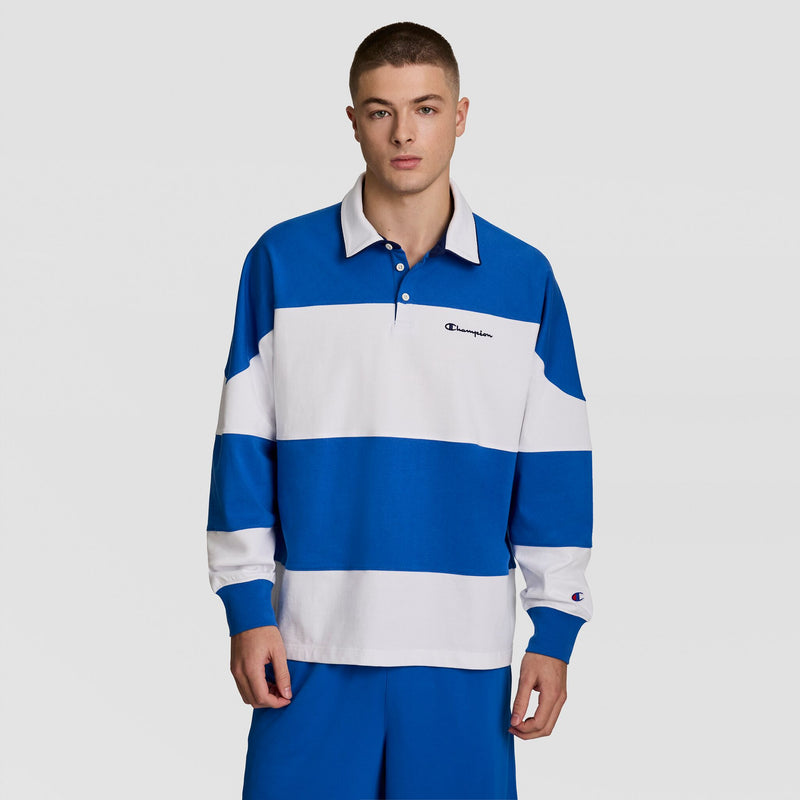 A person wears the Champion Gridiron Blocked Long Sleeve Rugby with a script logo and blue shorts, standing against a plain white background.