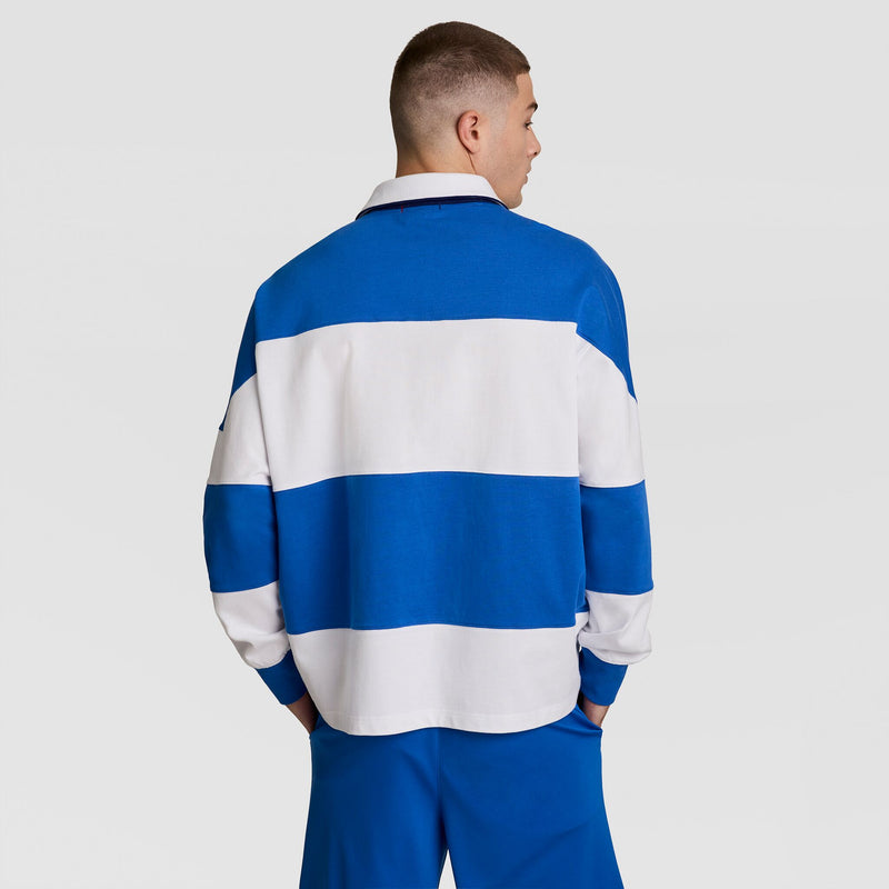 A person stands facing away, wearing the Champion Gridiron Blocked Long Sleeve Rugby with wide blue and white stripes and blue pants, against a plain light background.