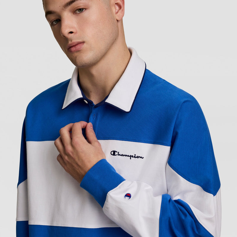 A person in a blue and white Champion Gridiron Blocked Long Sleeve Rugby with a white collar touches the cotton jersey near the embroidered script logo on the chest, set against a plain background.