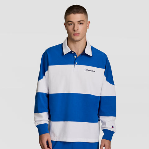 A young man wears the Champion Gridiron Blocked Long Sleeve Rugby with Script Logo, featuring blue and white stripes, a collared design, and soft cotton jersey, standing against a plain light background.