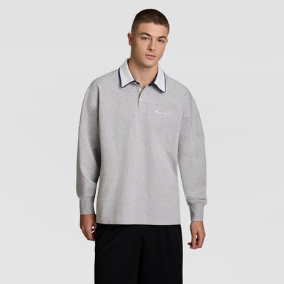 A young man wearing a light gray long-sleeve polo shirt and black shorts stands against a plain light gray background.