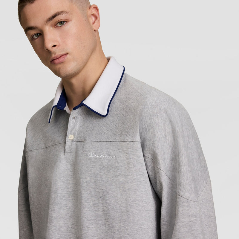 A young man with short hair wears a light gray long-sleeve polo shirt with a white and navy collar. The shirt has the Champion logo embroidered on the chest. The background is light gray.