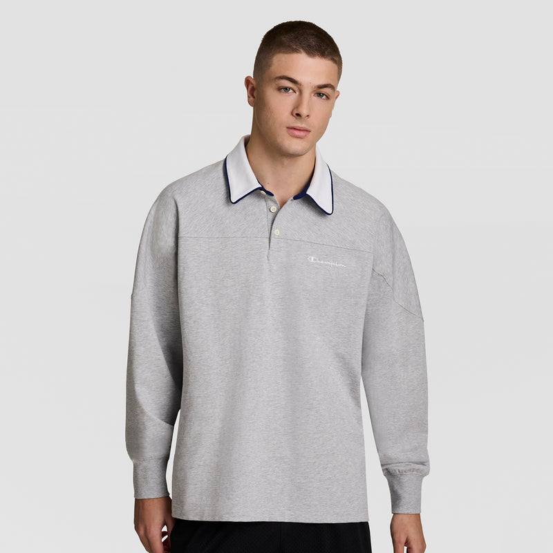 A young man with short hair wears the Champion Gridiron Long Sleeve Rugby, Script Logo, featuring a light gray design with a white and navy collar and the Champion logo on the left chest, standing against a plain background.