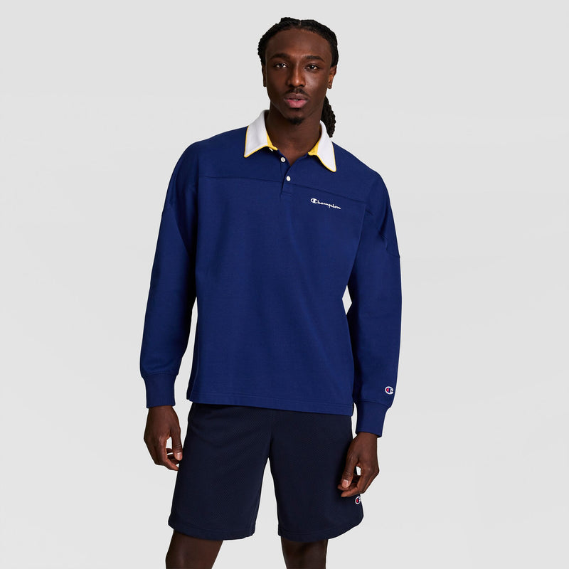 A man wears the Champion Gridiron Long Sleeve Rugby with a script logo—navy blue with a white collar—and matching navy shorts, standing against a plain light gray background.