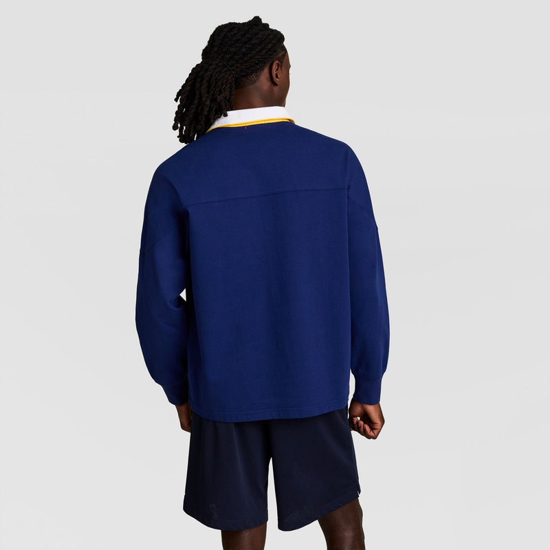 A person with long dark hair, wearing the Champion Gridiron Long Sleeve Rugby with Script Logo over a white collared shirt and navy shorts, stands facing away against a plain light background.