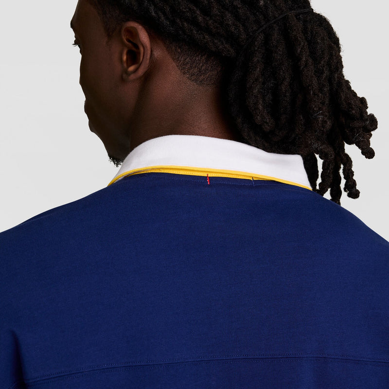 A person with dark skin and long, twisted hair pulled back is seen from behind, wearing the Champion Gridiron Long Sleeve Rugby with a white and yellow collar and script logo.