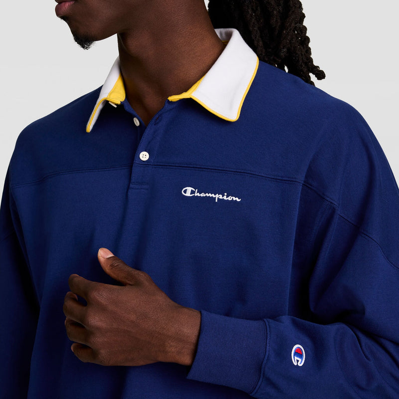 A person with dark skin and long hair wears the Champion Gridiron Long Sleeve Rugby shirt, featuring a blue design, yellow-trimmed white collar, and script logo embroidery on the chest and sleeve. They hold one hand to their chest.