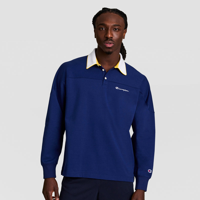 A man wears the Champion Gridiron Long Sleeve Rugby with Script Logo, featuring a navy blue design and white-yellow collar, standing against a light gray background. The shirt displays the iconic Champion logo on chest and sleeve.