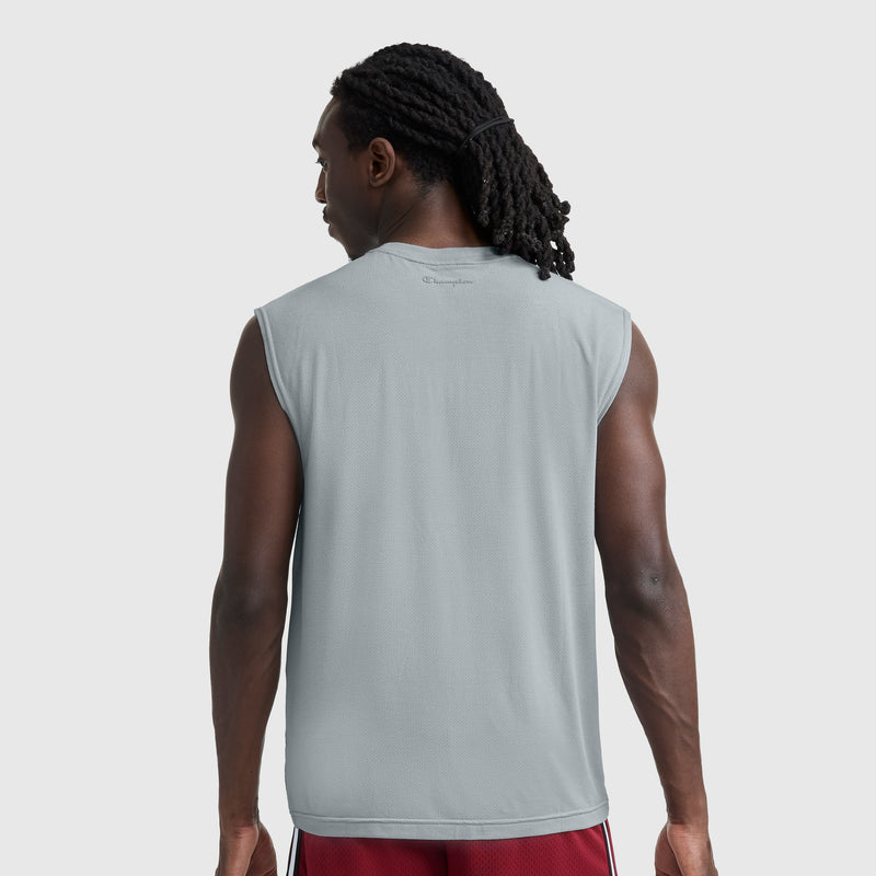 A man with long, dark, braided hair stands with his back to the camera against a plain light background, wearing a gray Champion Sport Crewneck Muscle Tee featuring the Script Logo and red shorts.