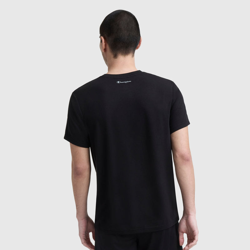 A person with short dark hair stands with their back to the camera, wearing a plain black Champion Sport Crewneck T-Shirt, Script Logo, and black shorts against a light gray background.