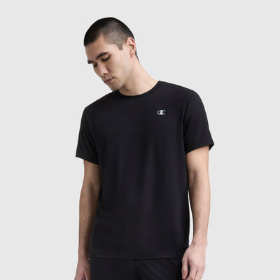 A young man with short dark hair wears a Champion Sport Crewneck T-Shirt, Script Logo, standing against a light gray background and looking down with a neutral expression.